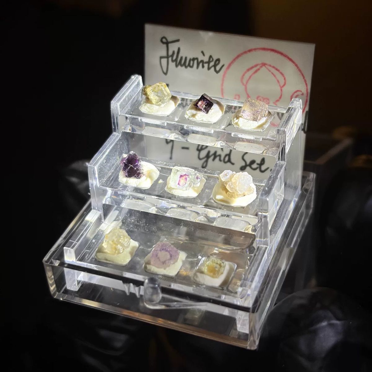 Set of nine mini fluorite crystals in a clear tiered display case, photo 1 of 3