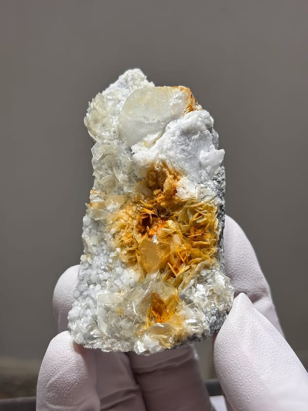 White feldspar plate with aquamarine, orange scheelite and mica from Xuebaoding, Sichuan, China, held in gloved hand