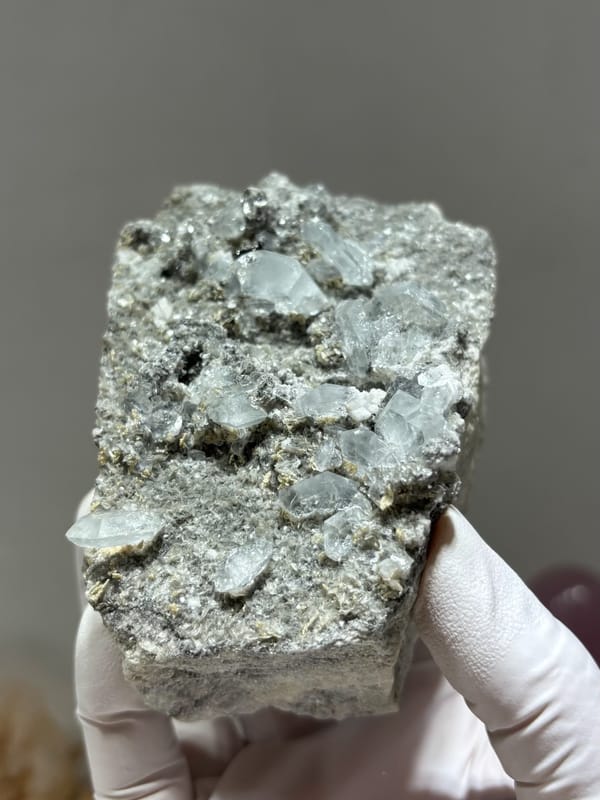 Grey matrix block dotted with pale aquamarine crystals from Xuebaoding, Sichuan, China, held in gloved hand