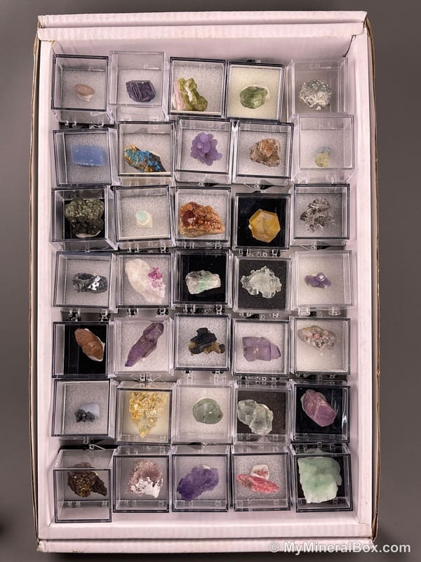 Bulk001 actual mixed mineral specimen box with about 35 different mini natural specimens in clear display boxes, including likely fluorite, quartz, aquamarine, pyrite, grape agate and other assorted minerals.