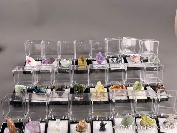 Bulk001 actual mixed mineral specimen box with about 35 different mini natural specimens in clear display boxes, including likely fluorite, quartz, aquamarine, pyrite, grape agate and other assorted minerals.