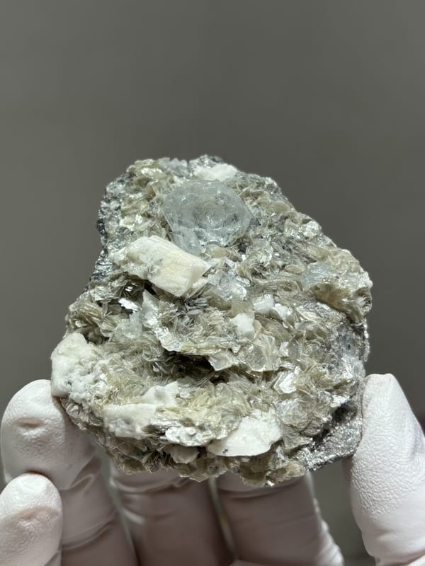 Pale aquamarine in grey mica with white feldspar from Xuebaoding, Sichuan, China, held in gloved hand