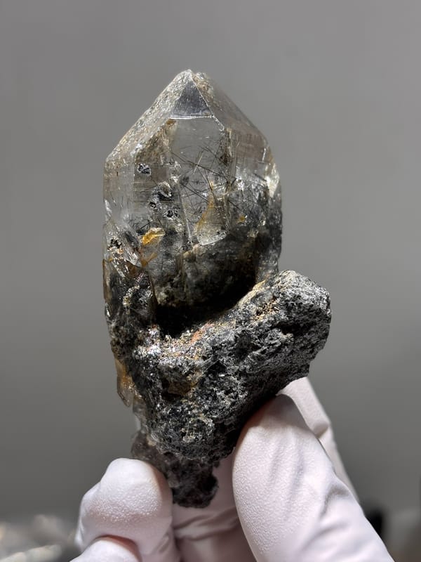 Smoky quartz point with black rutile on dark base from Xuebaoding, Sichuan, China, held in gloved hand