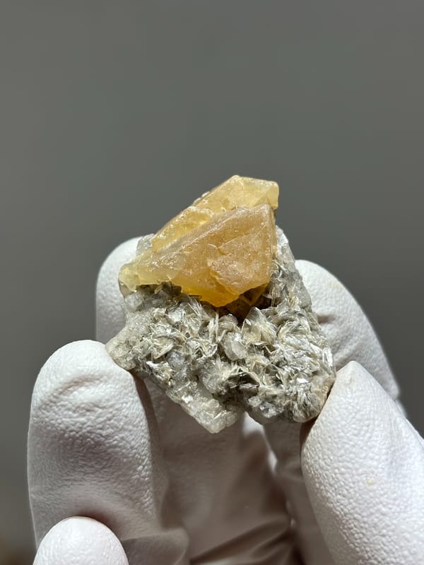 Honey scheelite crystal on mica with calcite base from Xuebaoding, Sichuan, China, held in gloved hand