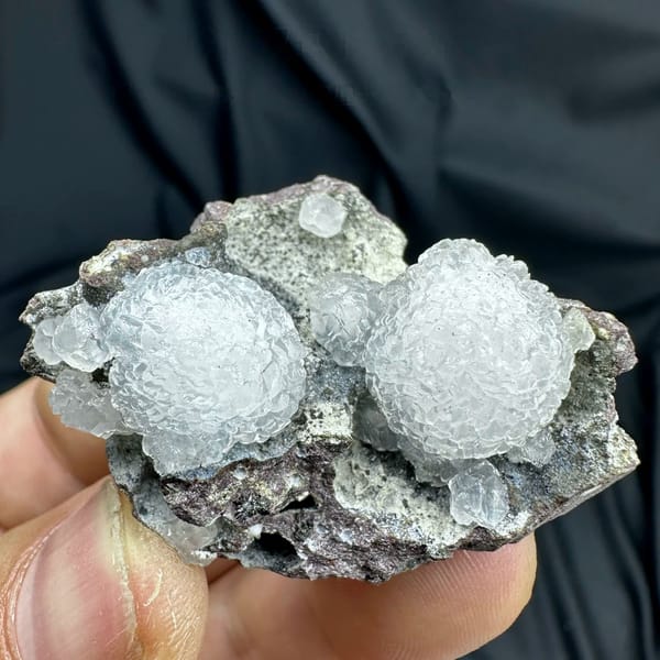 Two white botryoidal calcite spheres on grey matrix from Zhejiang, China, held in hand