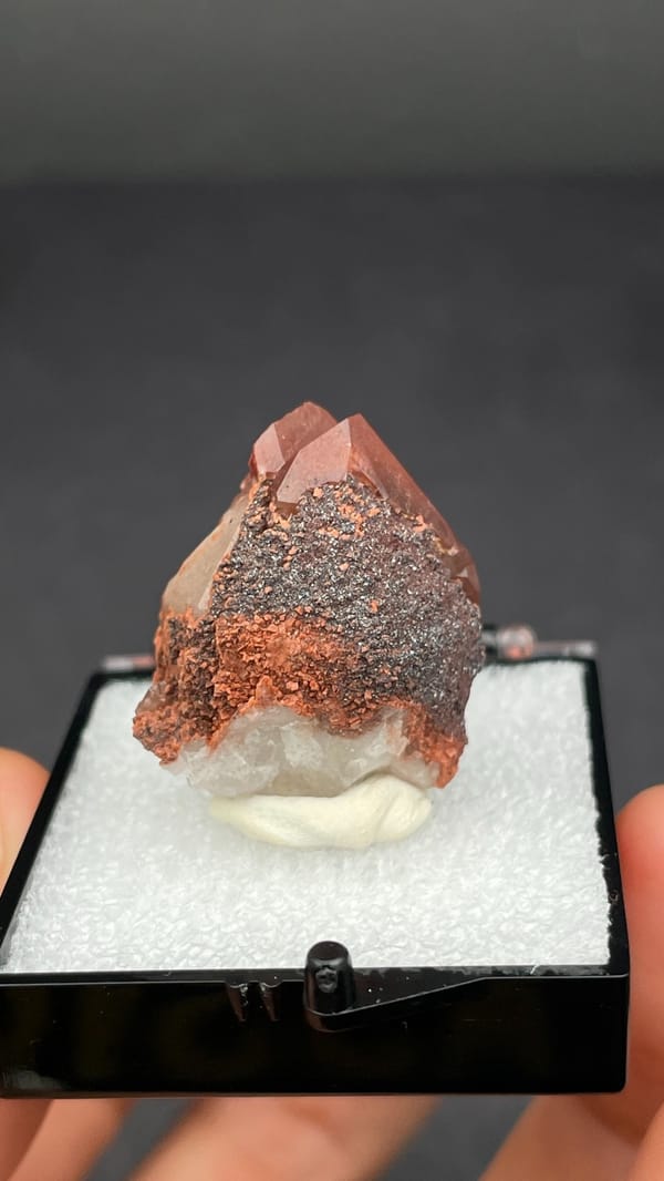 Pink-red quartz point with dark hematite crust from Shanxi, China, in a box, detail of the matrix contact
